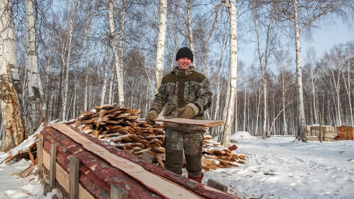 A man smiling while stacking wood in a snowy birch forest during winter.