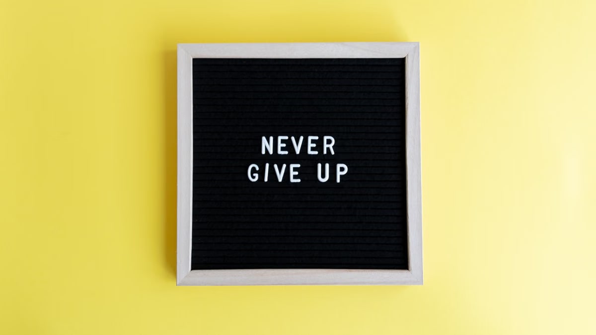 Black felt board with 'Never Give Up' on a bright yellow background for motivation.